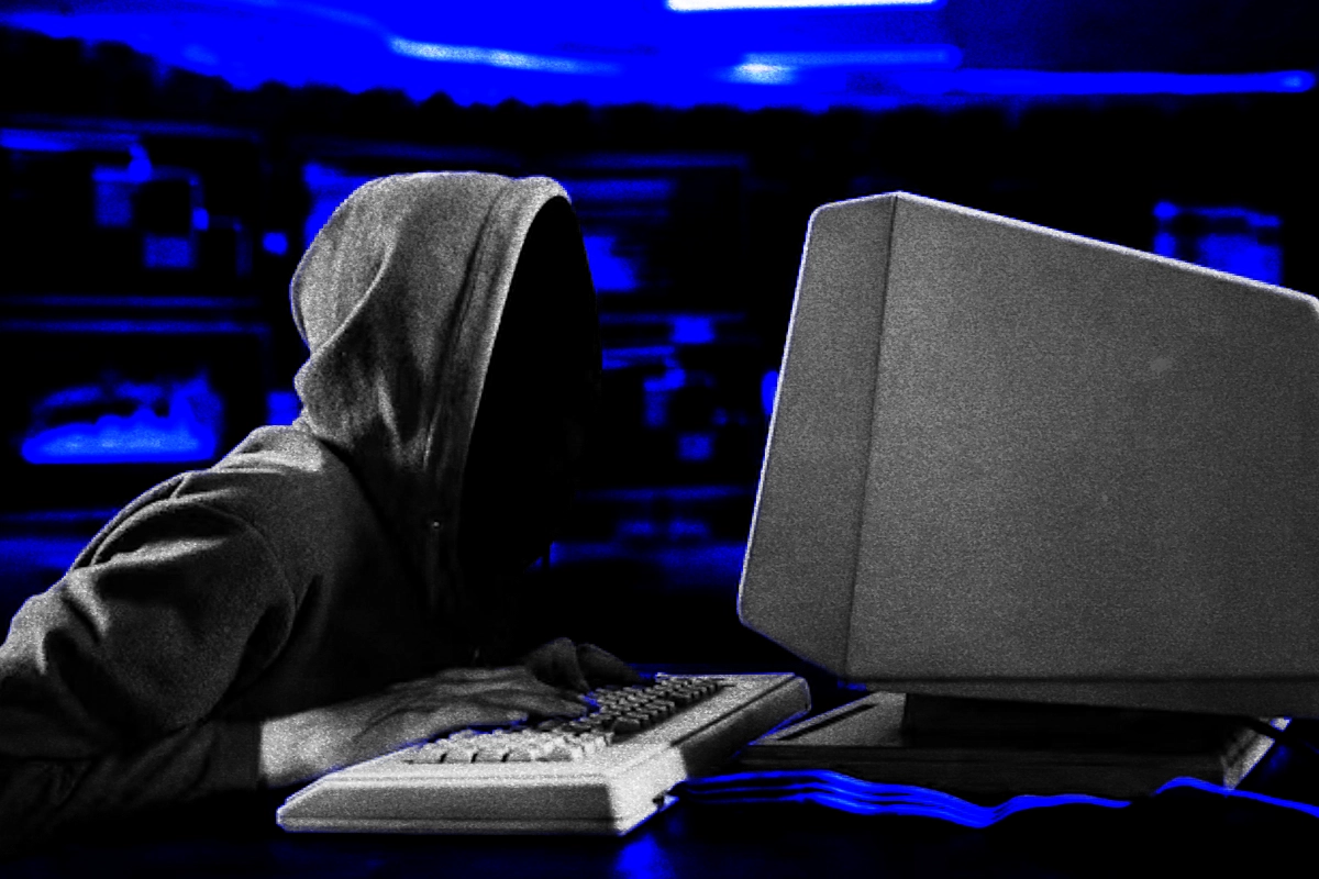 Hooded figure typing on a vintage keyboard before a bulky beige monitor, lit by blue glow