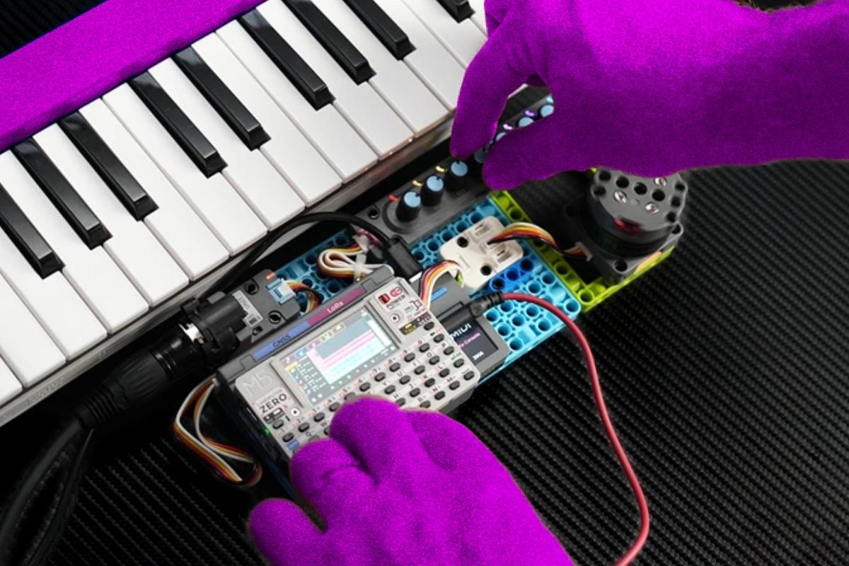 Purple-gloved hands operating a tiny keyboard computer wired into a synth rig with knobs
