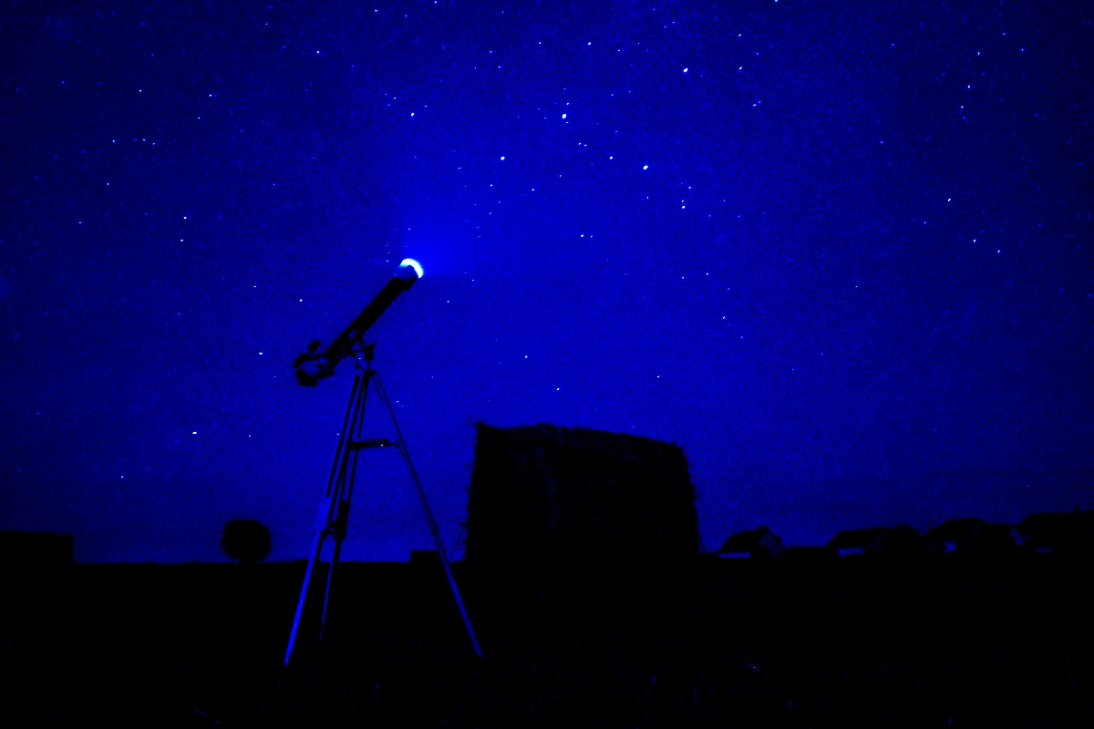 A telescope on a tripod points at a starry night sky beside a hay bale